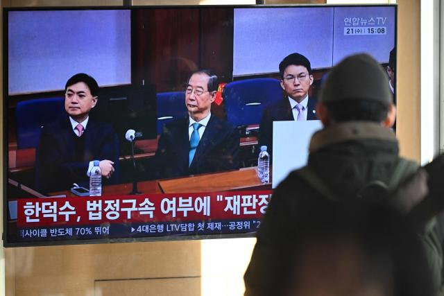 People watch a television screen showing a news broadcast of the trial verdict of South Korea's former prime minister Han Duck-soo (C on the screen) on charges related to martial law, at a train station in Seoul on January 21, 2026. A South Korean court sentenced former prime minister Han Duck-soo to 23 years in prison on January 21, for aiding and abetting a declaration of martial law that briefly suspended civilian rule. (Photo by Jung Yeon-je / AFP)