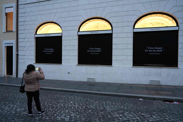 A woman takes a picture of Valentino's shop windows covered with black posters reading "I love beauty, It is not my fault - Valentino Garavani", in Rome on January 20, 2026. Valentino’s lying-in-state open today in Rome ahead of the funeral on January 23, 2026. (Photo by Stefano Costantino / AFP)