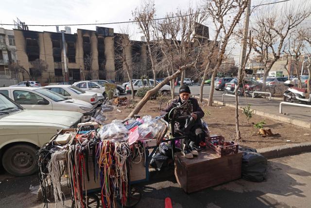 This photograph taken during a tour for foreign media shows a cobbler repairing shoes against the backdrop of government buildings that were burnt during recent public protests, in Tehran on January 21, 2026. Demonstrations sparked in late December by anger over economic hardship exploded into protests widely seen as the biggest challenge to the Iranian leadership in years. Limited internet access briefly returned in Iran before dropping again, a monitor said January 18, 10 days into a communications blackout that rights groups said aimed to mask a protest crackdown that killed thousands. (Photo by ATTA KENARE / AFP)