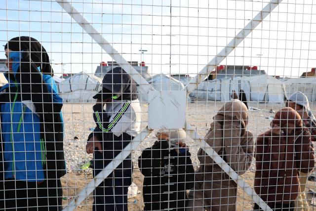 Young, relatives of suspected Islamic State jihadists, hide their faces as they stand behind a fence in Al-Hol camp in the desert region of Syria's Hasakeh province on January 21, 2026. Syria's army entered the vast Al-Hol camp that houses relatives of suspected Islamic State jihadists after Kurdish forces withdrew from the site, said an AFP journalist at the scene. The camp in a desert region of Hasakeh province holds around 24,000 people, including some 6,200 women and children from around 40 nationalities. (Photo by OMAR HAJ KADOUR / AFP)