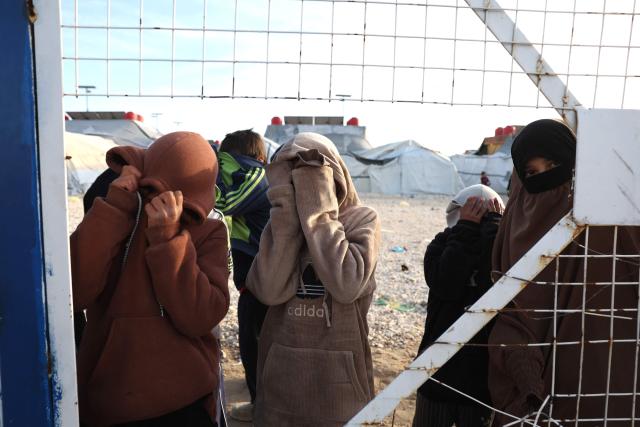 Young, relatives of suspected Islamic State jihadists, hide their faces as they stand behind a fence in Al-Hol camp in the desert region of Syria's Hasakeh province on January 21, 2026. Syria's army entered the vast Al-Hol camp that houses relatives of suspected Islamic State jihadists after Kurdish forces withdrew from the site, said an AFP journalist at the scene. The camp in a desert region of Hasakeh province holds around 24,000 people, including some 6,200 women and children from around 40 nationalities. (Photo by OMAR HAJ KADOUR / AFP)
