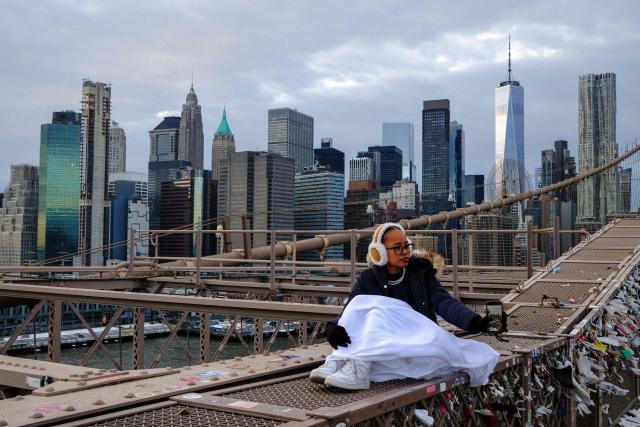 A woman records the city skyline with her phone, bracing the cold temperatures on the Brooklyn Bridge in the Manhattan borough of New York City on January 21, 2026. Bringing icy temperatures, Winter Storm Fern will slam a massive stretch of the United States this week, with more than 175 million people facing the prospect of heavy snowfall, power outages and travel disruption. Forecasters warned it could be 2,000 miles (3,219 kilometers) long -- well over half the length of the continental US. (Photo by CHARLY TRIBALLEAU / AFP)