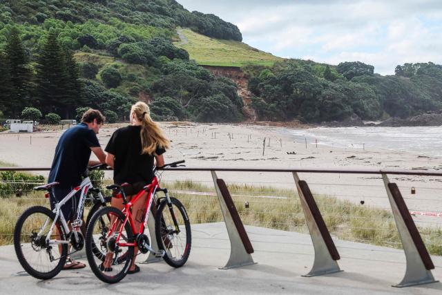 Cyclists stop to look at a landslide while a search is underway by local emergency services for missing people at Mount Maunganui in Tauranga on January 22, 2026. A landslide smashed into a campsite in rain-swept northern New Zealand leaving multiple people missing, police and rescuers said. (Photo by DJ MILLS / AFP)