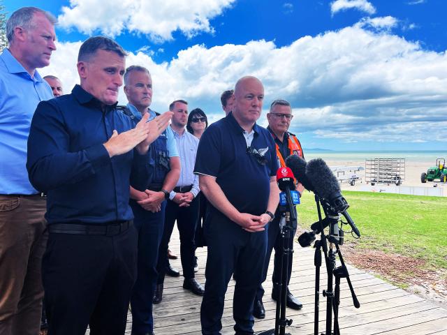 New Zealand Prime Minister Christopher Luxon (2nd-R) speaks to the media near the site of a landslide at Mount Maunganui in Tauranga on January 23, 2026. Rescuers dug into deep mud searching for at least six missing people on January 23, after a landslide from an extinct volcano crashed into a popular campsite in northern New Zealand. (Photo by Ben STRANG / AFP)