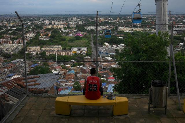 A man rests on a bench while cable cars from the "MIO Cable" public transport system cross the Siloe neighborhood in Cali, Colombia, on January 23, 2026. Santiago de Cali, often referred to as Cali, is the capital of Colombia's Valle del Cauca department and the country's third-largest city. (Photo by JOAQUIN SARMIENTO / AFP)