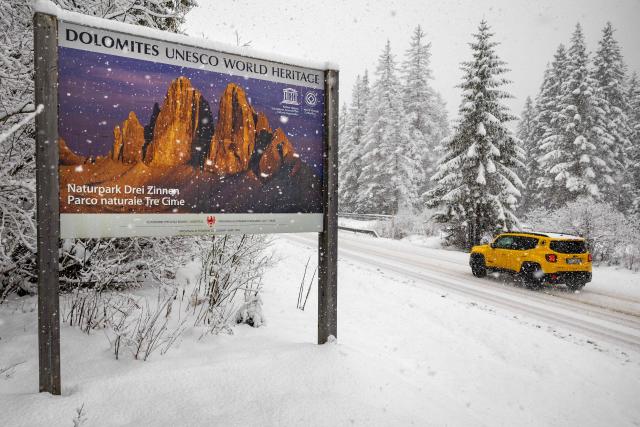 A motorist travels in heavy snow fall past a poster with an image of a sunnier day along a mountain pass road near Toblach running between the Olympic venues in Cortina and Antholz, northern Italy prior to the Milano Cortina 2026 Olympic Games, on January 25, 2026. (Photo by Odd ANDERSEN / AFP)