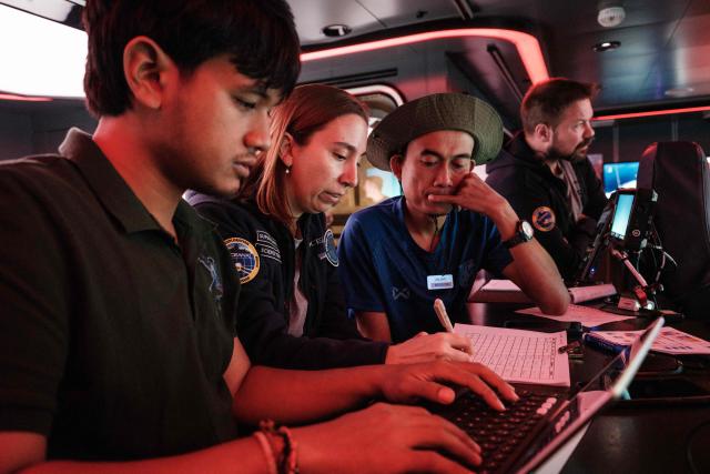 This photo taken on January 10, 2026, shows Larissa Fruehe (2L), a German environmental DNA (eDNA) scientist, speaking with Indonesian marine scientists as they monitor remotely operated vehicle (ROV) activity in the deep sea from a control room on the OceanXplorer, a research vessel operated by the marine non-profit OceanX, in waters off Sulawesi island. A dome-fronted submersible sinks beneath the waves off Indonesia, heading nearly 1,000 metres down to the realm of purple-frilled corals and bioluminescent shrimp. This month, AFP boarded one of two submersibles belonging to OceanX, a non-profit founded by billionaire Ray Dalio and his son that works with scientists on its OceanXplorer ship to study the marine world. (Photo by YASUYOSHI CHIBA / AFP) / To go with: INDONESIA-OCEANX-OCEANS-ENVIRONMENT-RESEARCH-SCIENCE, by Sara HUSSEIN