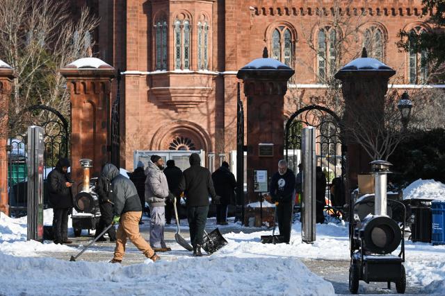 Workers clear snow from a sidewalk near the Smithsonian Insitution Building, also known as the Smithsonian Castle, along the National Mall in Washington, DC on January 26, 2026. A monster storm barreling across the United States had killed at least 11 people on Monday, prompting warnings to stay off the roads, mass flight cancelations and power outages after a weekend of misery. The storm dumped snow, sleet and freezing rain across swathes of the country from Texas to New England, with temperatures set to fall dangerously low this week. (Photo by Mandel NGAN / AFP)