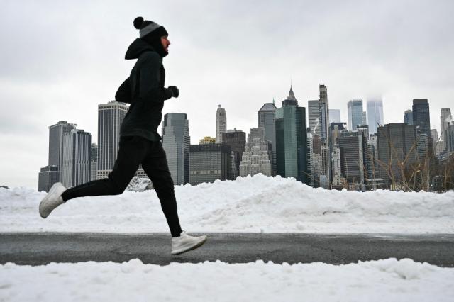 The Lower Manhattan skyline is seen in the background as a man runs along a path in the Brooklyn borough of New York City on January 26, 2026. A monster storm barreling across the United States had killed at least 11 people on Monday, prompting warnings to stay off the roads, mass flight cancelations and power outages after a weekend of misery. The storm dumped snow, sleet and freezing rain across swathes of the country from Texas to New England, with temperatures set to fall dangerously low this week. (Photo by ANGELA WEISS / AFP)