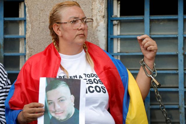 A woman chained to a railing holds a picture of her beloved one while waiting for news on the release of prisoners, outside of the Zone 7 of the Bolivarian National Police (PNB) in Sucre municipality, Metropolitan District of Caracas (DMC) on January 26, 2026. More than 100 political prisoners were freed on the eve in Venezuela, where detainees are slowly being released under pressure from the United States, the non-governmental organization Foro Penal said. (Photo by Pedro MATTEY / AFP)
