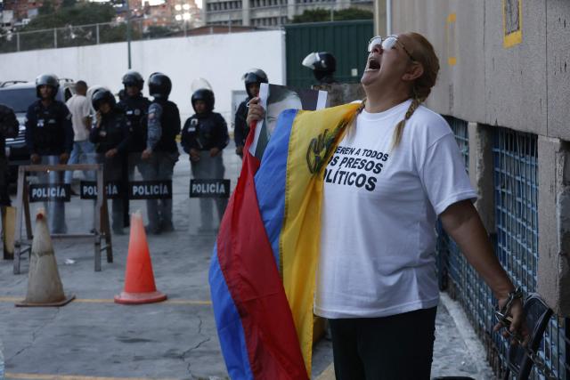 Evelis Cano, the mother of imprisoned Jack Tantak Cano, shouts slogans while holding a Venezuelan flag and chained to a railing as she waits for news about the release of prisoners outside Zone 7 of the Bolivarian National Police (PNB) in the Sucre municipality of the Metropolitan District of Caracas (DMC) on January 26, 2026. Evelis questions the slow process of political-prisoner releases announced on January 8 by Venezuelas interim government, under pressure from Washington after Nicolas Maduro was captured on January 3 in a US forces operation. (Photo by Pedro MATTEY / AFP)