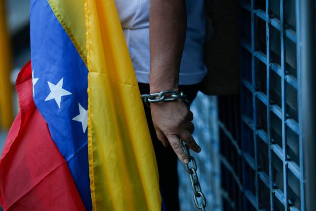This view shows the hand of Evelis Cano, the mother of imprisoned Jack Tantak Cano, chained to a railing and holding a Venezuelan flag as she waits for news about the release of prisoners outside Zone 7 of the Bolivarian National Police (PNB) in the Sucre municipality of the Metropolitan District of Caracas (DMC) on January 26, 2026. Evelis questions the slow process of political-prisoner releases announced on January 8 by Venezuelas interim government, under pressure from Washington after Nicolas Maduro was captured on January 3 in a US forces operation. (Photo by Pedro MATTEY / AFP)