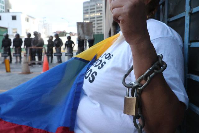 Evelis Cano, the mother of imprisoned Jack Tantak Cano, shouts slogans while holding a Venezuelan flag and chained to a railing as she waits for news about the release of prisoners outside Zone 7 of the Bolivarian National Police (PNB) in the Sucre municipality of the Metropolitan District of Caracas (DMC) on January 26, 2026. Evelis questions the slow process of political-prisoner releases announced on January 8 by Venezuelas interim government, under pressure from Washington after Nicolas Maduro was captured on January 3 in a US forces operation. (Photo by Pedro MATTEY / AFP)