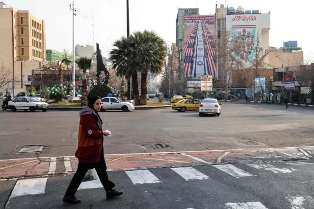 A woman crosses a street as cars drive past an anti-US and anti-Israel banner hanging on a building in Palestine Square in Tehran on January 27, 2026. A US naval strike group led by an aircraft carrier has deployed to Middle Eastern waters, the United States said on January 26, 2026, as Tehran warned it was ready to hit back at any American attack launched in response to a crackdown on anti-government protests. (Photo by ATTA KENARE / AFP)