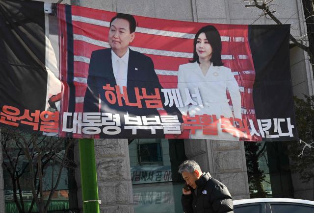 A man walks past a banner showing a picture of South Korea's former impeached president Yoon Suk Yeol and his wife Kim Keon Hee, on a street near the Seoul Central District Court in Seoul on January 28, 2026, as the banner reads "God will resurrect and restore Yoon Suk Yeol and his wife". A South Korean court will pass judgement on January 28, in the case of former first lady Kim Keon Hee, with prosecutors demanding a 15-year prison term over alleged stock fraud and corruption. (Photo by Jung Yeon-je / AFP)