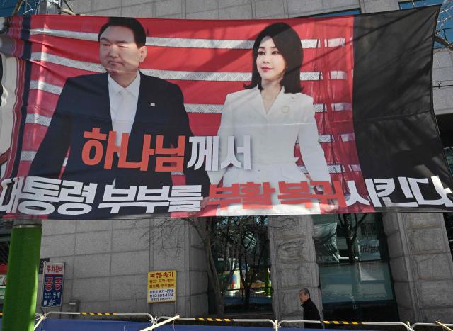 A man walks past a banner showing a picture of South Korea's former impeached president Yoon Suk Yeol and his wife Kim Keon Hee, on a street near the Seoul Central District Court in Seoul on January 28, 2026, as the banner reads "God will resurrect and restore Yoon Suk Yeol and his wife". A South Korean court will pass judgement on January 28, in the case of former first lady Kim Keon Hee, with prosecutors demanding a 15-year prison term over alleged stock fraud and corruption. (Photo by Jung Yeon-je / AFP)