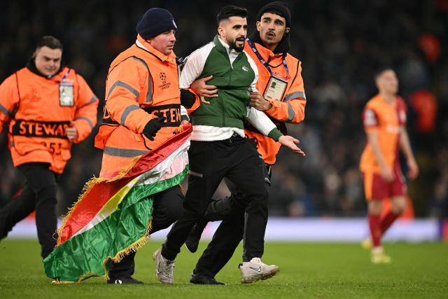 Am pitch invader carries a flag as they are escorted off the pitch during the UEFA Champions League football match between Manchester City and Galatasaray at the Etihad Stadium in Manchester, north west England, on January 28, 2026. (Photo by Oli SCARFF / AFP)