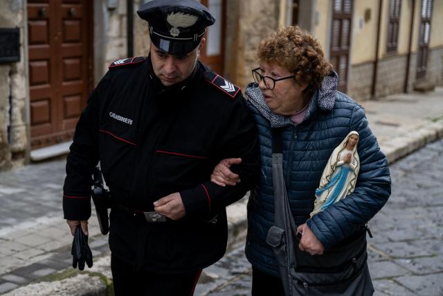 A woman helped by an Italian Carabinieri holds a statue of the Virgin Mary as she leaves her home after returning with firefighters to collect her belongings in the red zone of Niscemi, Sicily, on January 29, 2026. A large area of the town of Niscemi is declared  "red zone" after the a 4km-long stretch of the hillside collapsed on January 25, 2026, forcing the evacuation of some 1,500 people. The town, built on unstable terrain, was battered by a powerful storm which hit southern Italy last week and experts say the chasm could widen further and swallow houses with the rains expected over the coming days. (Photo by MARCO BERTORELLO / AFP)