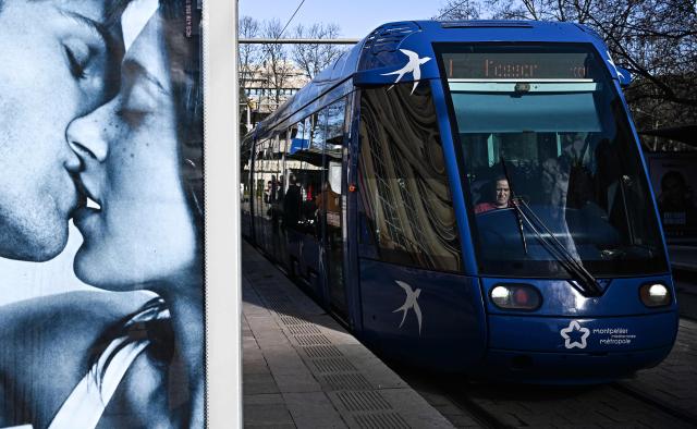 An employee drives a tramway past an advertisement placard depicting a couple kissing in Montpellier, southern France, on January 29, 2026. Free transportation is a popular idea for the March municipal elections in many cities. But the promises vary greatly, and some candidates denounce it as “vote fishing” or even a “false good idea.” Initially limited to young people and seniors, then to weekends, this promise made in 2020 for Montpellier has, for a little over two years now, been available on buses and trams to all 500,000 residents of this metropolitan area. (Photo by Gabriel BOUYS / AFP)