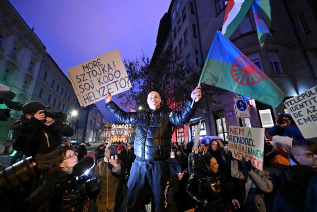 A man holds a flag of the Romani people and a placard reading  'Hey you Sztojka (Attila Sztojka, representative of the Hungarian Parliament and state secretary of Prime Minister Orban's government)! What's the situation now?' near parliament building as Roma activists and sympathisers take part in a peaceful demonstration in downtown Budapest on January 31, 2026. The protest was sparked by an earlier statement by Minister János Lázár, in which he suggested that tasks such as cleaning railway toilets should be performed by Roma people. Organisers stated that the aim of the demonstration was to protect the dignity of the Roma. (Photo by Attila KISBENEDEK / AFP)