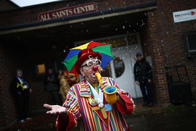 A clown performs in the rain on the way to attend the annual Grimaldi Memorial Service at the All Saints Church in east London on February 1, 2026. The annual event is open to clowns from across the world who gather in memory of the legendary Joseph Grimaldi, the "king of clowns", who was a 19th century English stage performer. (Photo by Henry NICHOLLS / AFP)