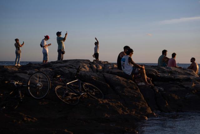 People enjoy the sunset while standing and sitting on rocks along the coast of Montevideo on February 1, 2026. (Photo by Eitan ABRAMOVICH / AFP)