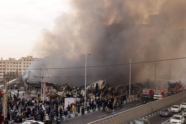 People stand and watch as firefighters battle a fire that broke out in Jannat Bazaar, west of Tehran on February 3, 2026. A massive fire broke out on February 3 at a bazaar west of the Iranian capital, state media reported, although the cause of the blaze was unclear. The fire erupted at a market in the Jannat Abad neighbourhood in Tehran's west, an area filled with stalls and shops, state TV quoted a spokesman for Tehran's fire department as saying. (Photo by ATTA KENARE / AFP)