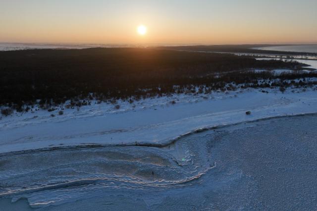 This aerial photograph taken on February 3, 2026 shows people walking along the Baltic Sea beach covered with ice thrown up by the waves as round ice floes float near Mikoszewo, northern Poland, with a winter low sunset sun in the background. (Photo by Sergei GAPON / AFP)