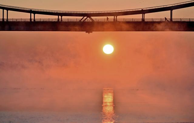 People walk on the pedestrian bridge over the Dnipro River during sunrise in Kyiv on February 4, 2026. (Photo by Sergei SUPINSKY / AFP)