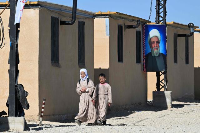 Girls walk near a poster of Lebanese Shiite movement Hezbollah's leader Naim Qassem displayed outside shelters at the Imam Ali Housing Compound, where displaced Lebanese and Syrian refugee Shiite Muslims take refuge by the city of Hermel in Lebanon's northeastern Bekaa valley on February 4, 2026. (Photo by Joseph EID / AFP)