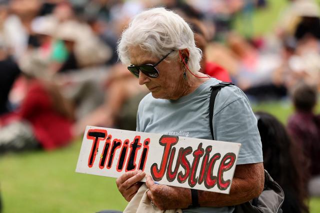 A protestor holds a placard during a ceremony to commemorate Waitangi Day in Waitangi on February 5, 2026. New Zealand's Prime Minister Christopher Luxon was heckled by a Maori crowd on February 5 during a speech marking national Waitangi Day celebrations, an annual political gathering that gives Indigenous tribes a chance to air grievances. (Photo by Ben STRANG / AFP)
