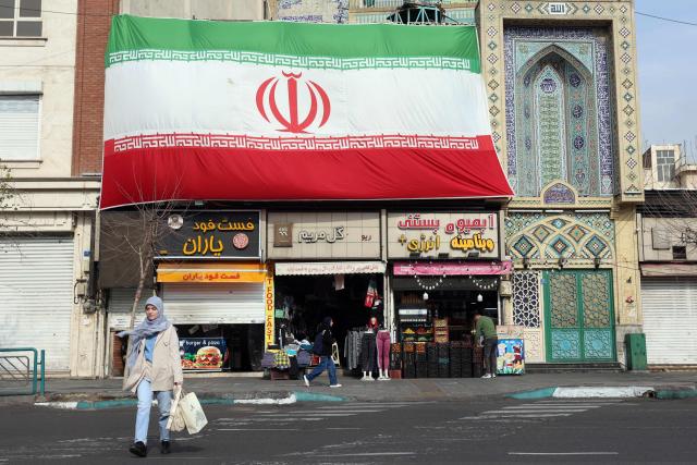 An Iranian woman walks past a huge national flag hanging above shops, in Tehran on February 6, 2026. Iran's foreign minister met with his Omani counterpart in Muscat on February 6, ahead of Oman-mediated talks with the United States on the Islamic republic's nuclear programme. (Photo by AFP)