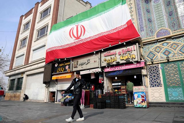 An Iranian man speaks on his mobile phone as he walks past a huge national flag hanging above shops, in Tehran on February 6, 2026. Iran's foreign minister met with his Omani counterpart in Muscat on February 6, ahead of Oman-mediated talks with the United States on the Islamic republic's nuclear programme. (Photo by AFP)