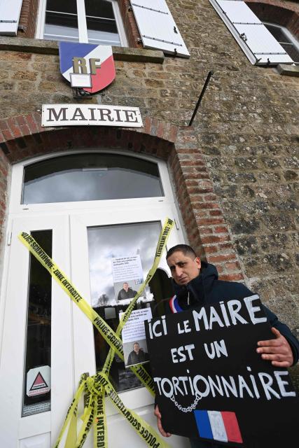 Member of the children's rights organization Mouv'Enfants Samir Baroualia holds a placard reading "Here the mayor is a torturer" as he demonstrates in front of the town hall to protest incumbent mayor Edgar Moulin's candidacy for re-election in Saint-Ellier-les-Bois, near Alencon, on February 6, 2026. The mayor has decided to maintain his candidacy despite being charged with rape of a person engaged in prostitution, aggravated pimping, soliciting minors for prostitution, and possession of firearms, and having spent several months in pretrial detention. (Photo by JEAN-FRANCOIS MONIER / AFP)