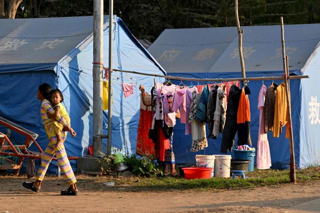 A woman, displaced after clashes between Thai and Cambodian soldiers, carries a child past tents in an evacuation centre on the grounds of a pagoda in Banteay Meanchey province on February 4, 2026. A sign hanging from a rusty ice-green shipping container installed by Thai forces on what they say is the border with Cambodia proclaims: "Cambodian citizens are strictly prohibited from entering this area." (Photo by TANG CHHIN Sothy / AFP) / To go with 'THAILAND-CAMBODIA-CONFLICT-BORDER,REPORTAGE' by Suy SE and Montira RUNGJIRAJITTRANON