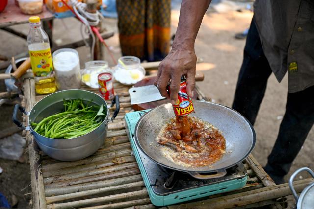 Farmer Sok Chork, displaced after clashes between Thai and Cambodian soldiers, cooks a dish from canned sardines in front of his shelter in an evacuation centre on the grounds of a pagoda in Banteay Meanchey province on February 4, 2026. A sign hanging from a rusty ice-green shipping container installed by Thai forces on what they say is the border with Cambodia proclaims: "Cambodian citizens are strictly prohibited from entering this area." (Photo by TANG CHHIN Sothy / AFP) / To go with 'THAILAND-CAMBODIA-CONFLICT-BORDER,REPORTAGE' by Suy SE and Montira RUNGJIRAJITTRANON