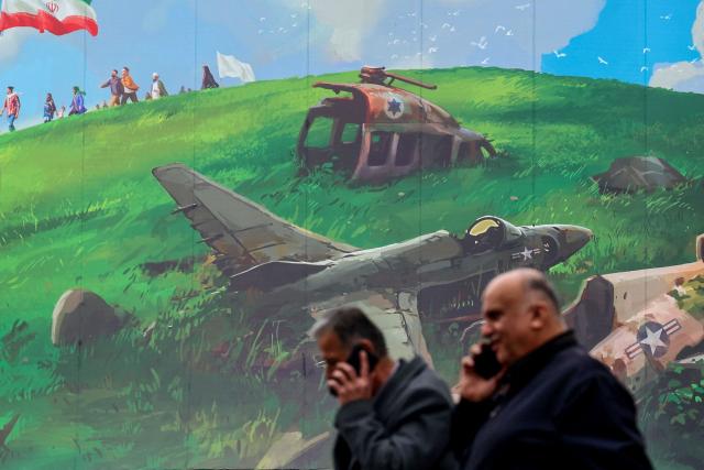 Iranian men speaking on their mobile walk past a huge anti-US billboard displayed on a building in Tehran's Valiasr Square on February 8, 2026. (Photo by ATTA KENARE / AFP)