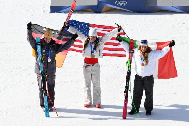 (From L) Silver medallist Germany's Emma Aicher, Gold medallist US' Breezy Johnson and bronze medallist Italy's Sofia Goggiaon pose with their national flags during the podium ceremony of the women's downhill event during the Milano Cortina 2026 Winter Olympic Games at the Tofane Alpine Skiing Centre in Cortina d’Ampezzo on February 8, 2026.  (Photo by Marco BERTORELLO / AFP)