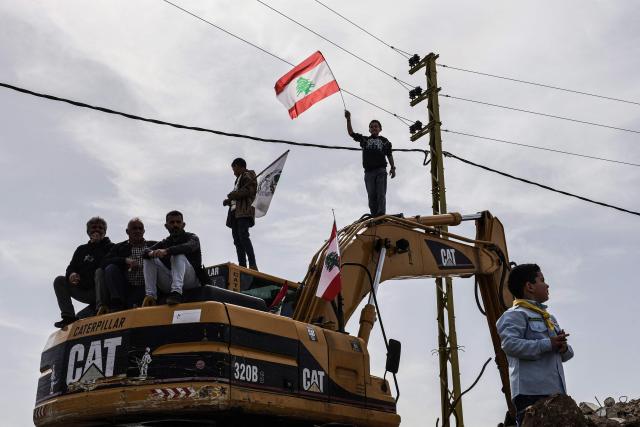 Standing on a digger used to clear the rubble of destroyed homes abd businesses, a Lebanese youth holds up the Lebanese flag as residents wait for the arrival of Lebanon's prime minister during his visit to the heavily-damaged southern village of Kfar Shouba, near the border with Israel on February 8, 2026. Swathes of south Lebanon's border areas remain in ruins and largely deserted more than a year after a US-brokered November 2024 ceasefire sought to end hostilities between Israel and the Iran-backed group. Salam vowed authorities would begin key projects including restoring roads, communications networks and water in the two towns (Photo by Rabih DAHER / AFP)