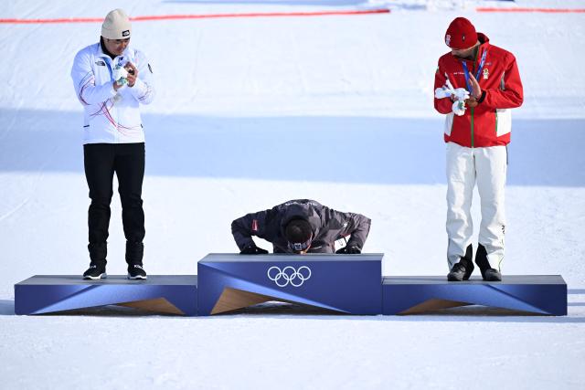 Silver medallist South Korea's Kim Sangkyum (L) and bronze-medallist Bulgaria's Tervel Zamfirov (R) look on as gold medallist Austria's Benjamin Karl (C) kisses the podium after the snowboard men's parallel giant slalom final at Livigno Snow Park during the Milano Cortina 2026 Winter Olympic Games, in Livigno (Valtellina), on February 8, 2026. (Photo by Kirill KUDRYAVTSEV / AFP)