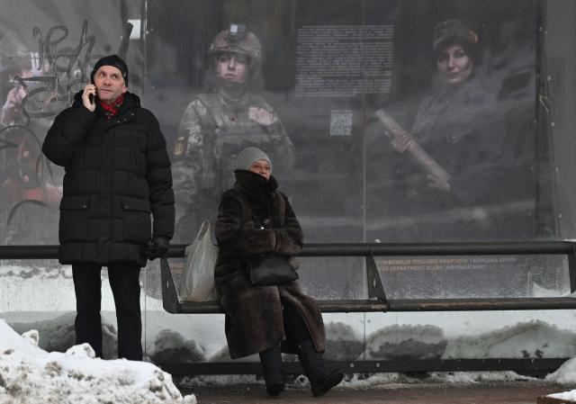 People wait at a bus stop in front of posters, featuring soldiers, displayed by the National Institute of National Remembrance as part of an open air exhibition in the center of Kyiv on February 8, 2026, amid the Russian invasion in Ukraine. (Photo by Sergei SUPINSKY / AFP)