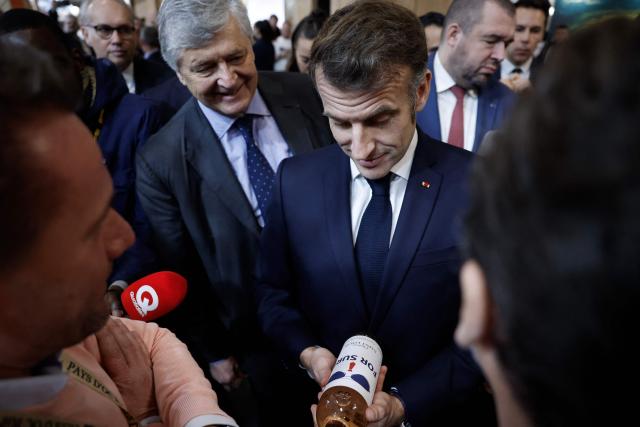 French President Emmanuel Macron (C-R) holds a bottle of wine "For Sure" next to French junior Minister in charge of external trade Nicolas Forissier as he visits the Wine Paris 2026 fair at the Porte de Versailles exhibition centre in Paris on February 9, 2026. Macron's mangling of "for sure" to "fo shur" in his Davos speech as he stood up to US president wearing his now iconic sunglasses also went viral from India to Indiana." (Photo by Yoan VALAT / POOL / AFP)