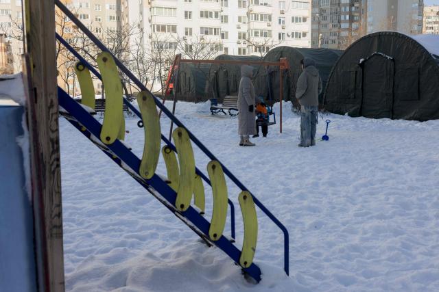Local residents play with a child on a swing set near emergency service tents set up for those whose homes are without electricity or heating following Russian missile and drone attacks on Ukrainian energy infrastructure, in a residential neighbourhood of Kyiv on February 10, 2026, amid the Russian invasion of Ukraine. (Photo by Tetiana DZHAFAROVA / AFP)