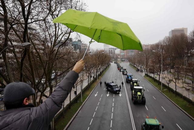 A man waves his umbrella to Spanish farmers protesting with tractors against EU-Mercosur trade deal and the economic pressures facing the agricultural sector, in Madrid on February 11, 2026. Spain on January 9 celebrated the approval by EU nations of a vast trade deal with South American bloc Mercosur, championed by business groups but loathed by many European farmers. (Photo by Oscar DEL POZO / AFP)
