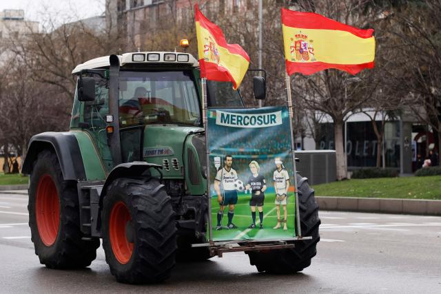 A poster placed on a tractor shows a caricature of European Commission President Ursula von der Leyen refereeing a football match between American and European farmers as Spanish farmers protest with tractors against EU-Mercosur trade deal and the economic pressures facing the agricultural sector, in Madrid on February 11, 2026. Spain on January 9, 2026 celebrated the approval by EU nations of a vast trade deal with South American bloc Mercosur, championed by business groups but loathed by many European farmers. (Photo by Oscar DEL POZO / AFP)