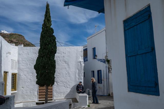 This photograph taken on February 4, 2026 shows woman talking with an eldery man in the village of Plaka in the Greek Aegean island of Milos. On the sloping coastline facing the Aegean Sea, the vast construction site looks like a gaping wound inflicted on the volcanic rock. ‘An ecological crime’ according to Manolis Mikelis, mayor of Milos, the Greek island in the Cyclades where this luxury hotel has begun extensive expansion work. "The geological uniqueness of Milos is known throughout the world. We don't want its identity to change," grumbles the mayor. Private villas and high-end complexes: the entire archipelago, known for its whitewashed houses and blue church domes, is caught up in a real estate fever fuelled by the tourism boom. (Photo by Aris MESSINIS / AFP)
