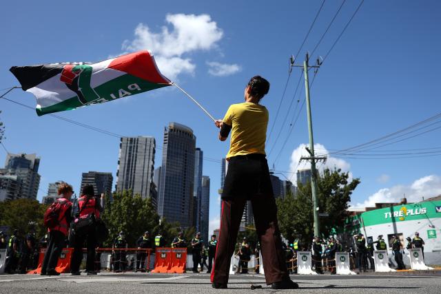 A protester waves a flag in front of police officers as they cordon off a venue during the visit of Israel’s President Isaac Herzog in Melbourne on February 12, 2026. Herzog's tightly secured, four-day trip aims to console Australia's Jewish community after the December shooting at Sydney's Bondi Beach that killed 15 people at a Hanukkah festival. (Photo by MARTIN KEEP / AFP)