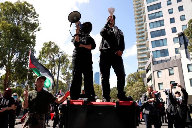 Two protesters stand atop a rubbish bin as they speak through megaphones in front of police officers as they cordon off a venue during the visit of Israel’s President Isaac Herzog in Melbourne on February 12, 2026. Herzog's tightly secured, four-day trip aims to console Australia's Jewish community after the December shooting at Sydney's Bondi Beach that killed 15 people at a Hanukkah festival. (Photo by MARTIN KEEP / AFP)