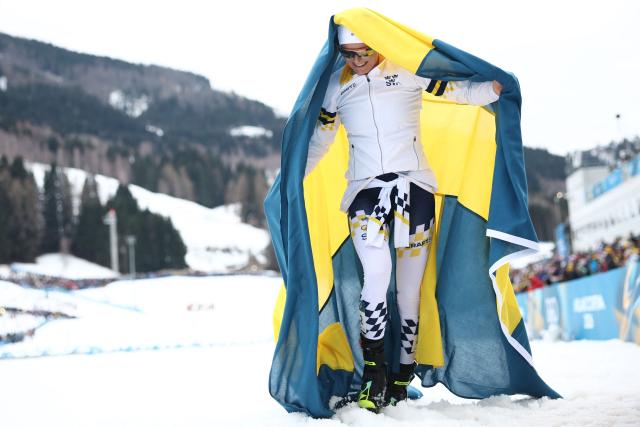 Sweden's Frida Karlsson starts to celebrate with Sweden's flag as the race is not over yet, during the women's cross-country skiing 10km interval start free event of the Milano Cortina 2026 Winter Olympic Games at Tesero Cross-Country Skiing Stadium in Lago di Tesero (Val di Fiemme), on February 12, 2026. (Photo by Anne-Christine POUJOULAT / AFP)