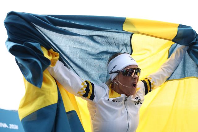 Sweden's Frida Karlsson starts to celebrate with Sweden's flag as the race is not over yet, during the women's cross-country skiing 10km interval start free event of the Milano Cortina 2026 Winter Olympic Games at Tesero Cross-Country Skiing Stadium in Lago di Tesero (Val di Fiemme), on February 12, 2026. (Photo by Anne-Christine POUJOULAT / AFP)